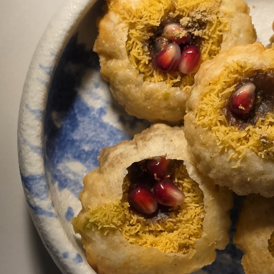 Crispy puris filled with spiced chutney, sev, and pomegranate seeds on a blue-patterned plate.